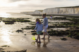 Two people walking with a small yellow net, on wet sand. Cliffs, rocks and sea can be seen in background.