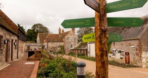 Farm buildings and courtyard in background. Signpost pointing in different directions to Eastbourne, Winchester, Lewes and Southease in foreground.